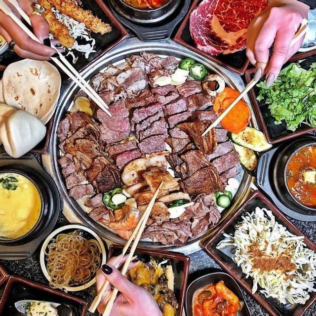 Una vista dall'alto di un abbondante piatto di barbecue coreano con diverse carni e verdure, circondato da contorni, mentre le persone mangiano con le bacchette.