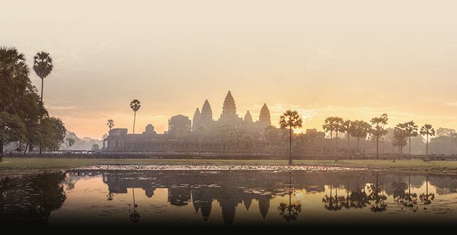The Angkor Wat temple silhouetted against a golden sunrise, its form reflected in a tranquil pond surrounded by palm trees.