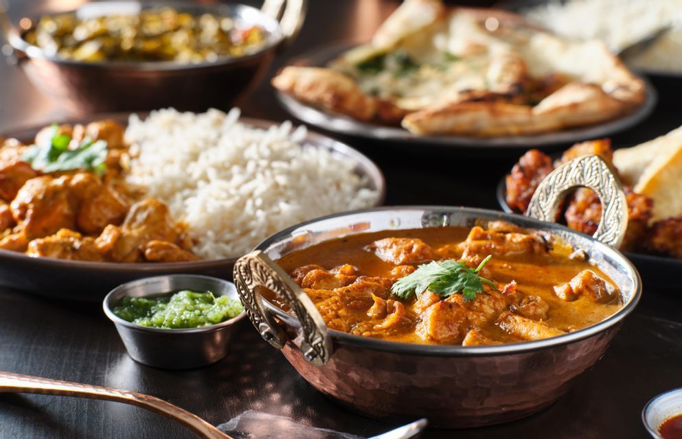 Un primo piano di una selezione di cibo indiano, con pollo al curry in una ciotola di rame, riso, chutney e pane naan sullo sfondo.