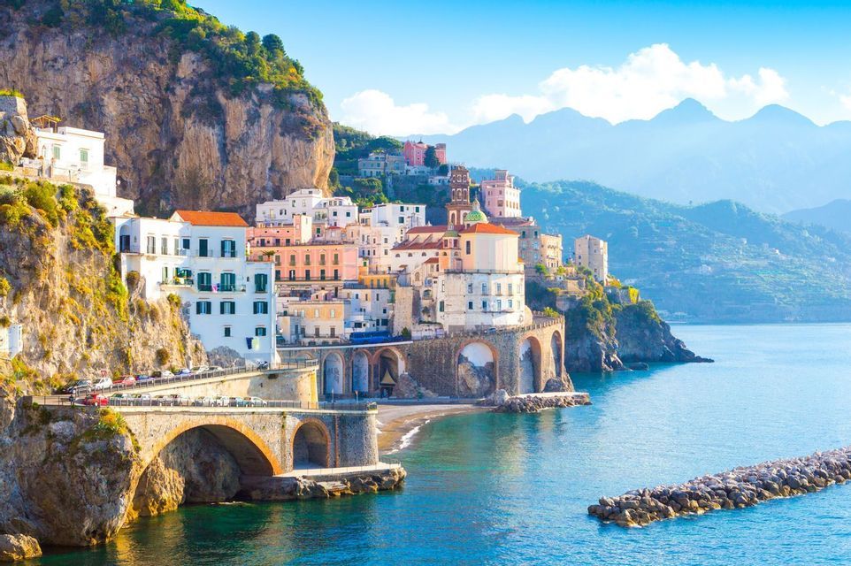 Eine Küstenstadt mit farbenfrohen Häusern an einer steilen Klippe und einer steinernen Bogenbrücke über dem blauen Meer.