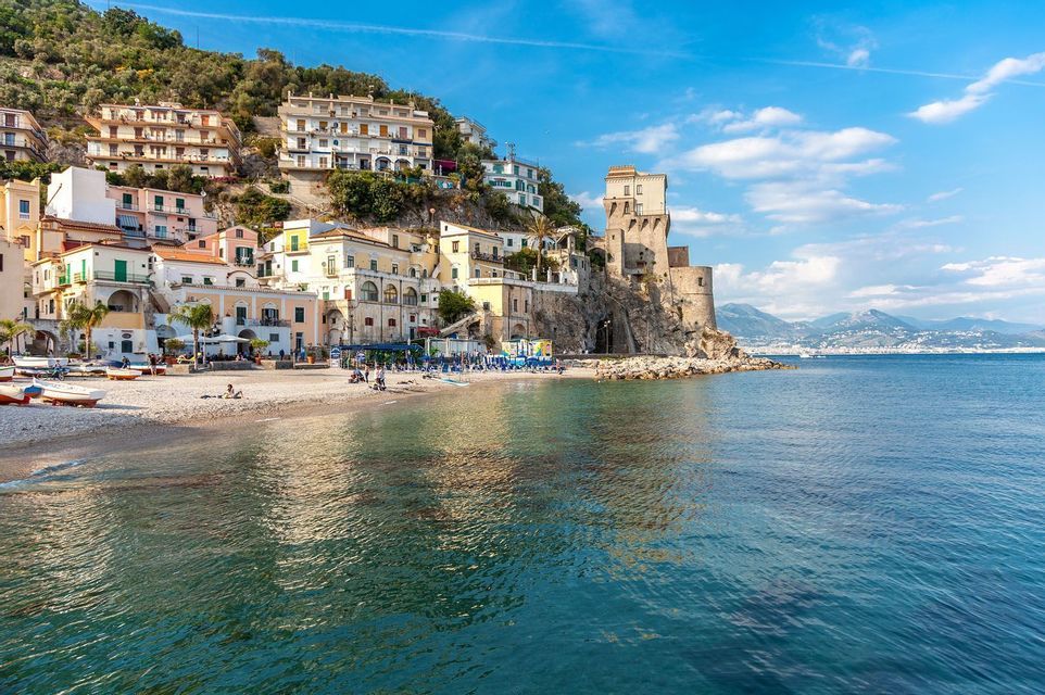 A coastal village with a watchtower and beach, viewed from the water under a blue sky with light clouds.