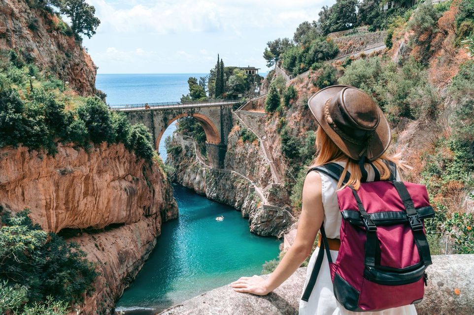 Une femme avec un chapeau et un sac à dos contemple des gorges profondes avec de l'eau turquoise, un pont de pierre et la mer en arrière-plan.