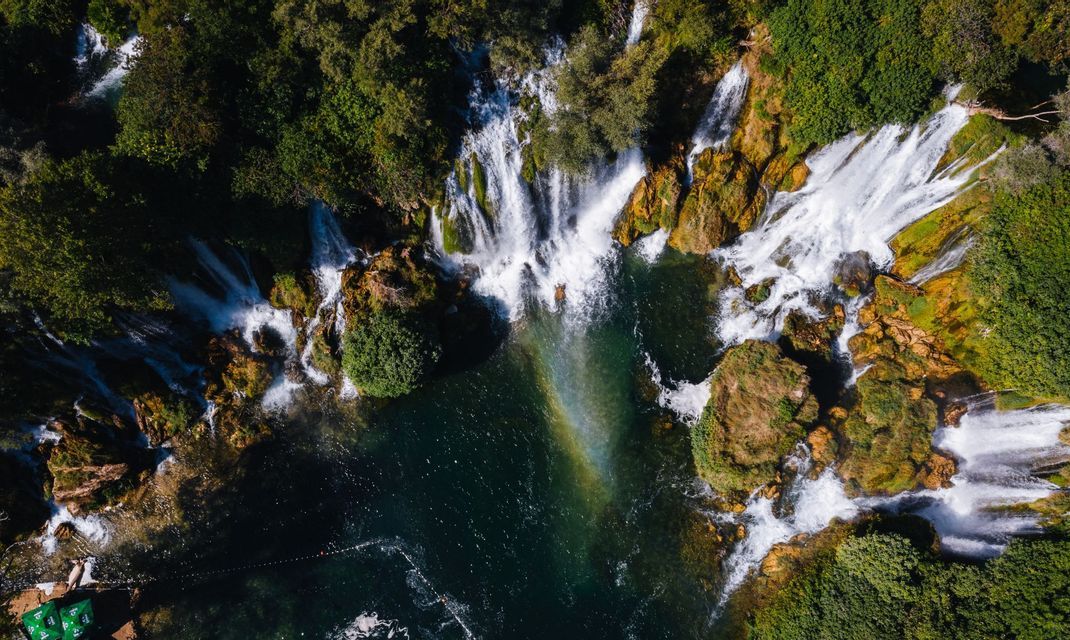 Vista aerea dall'alto di cascate che si gettano su rocce muschiose e alberi in una pozza turchese, con un arcobaleno nella nebbia.