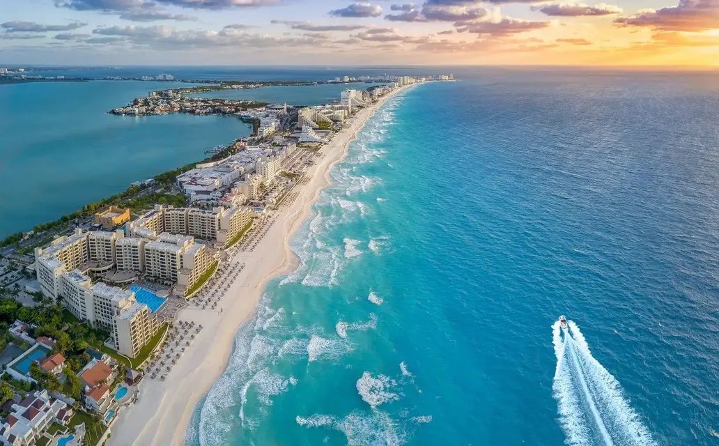 Veduta aerea di una costa con hotel lungo una spiaggia di sabbia bianca bagnata da acque turchesi, mentre un motoscafo sfreccia al tramonto.