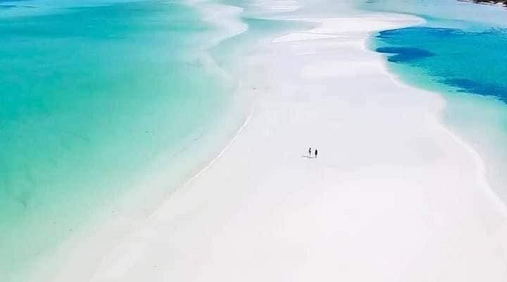 Una vista aerea di due persone in piedi su un vasto banco di sabbia bianca circondato da acque cristalline turchesi.