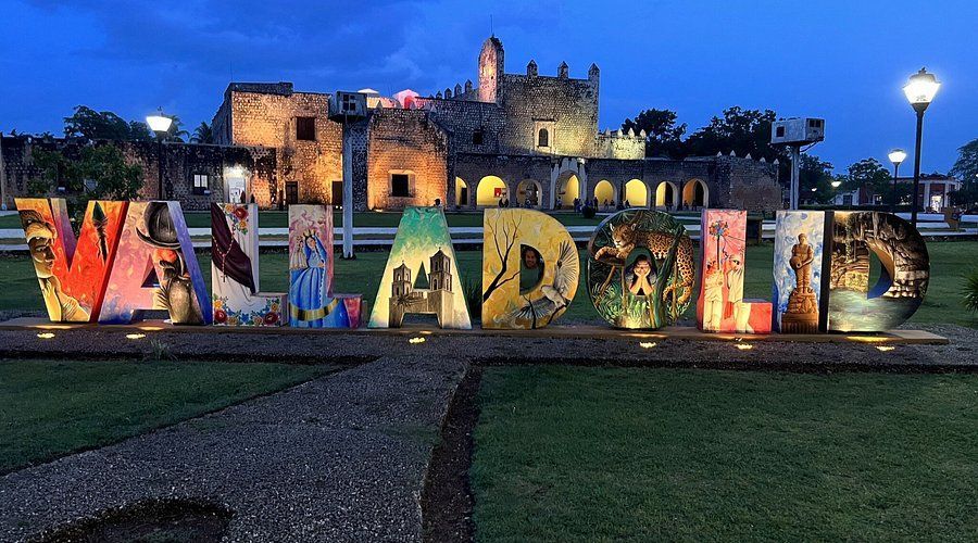 Lettere illuminate e colorate che compongono 'VALLADOLID' su un prato verde, con un vecchio edificio in pietra illuminato sullo sfondo al tramonto.