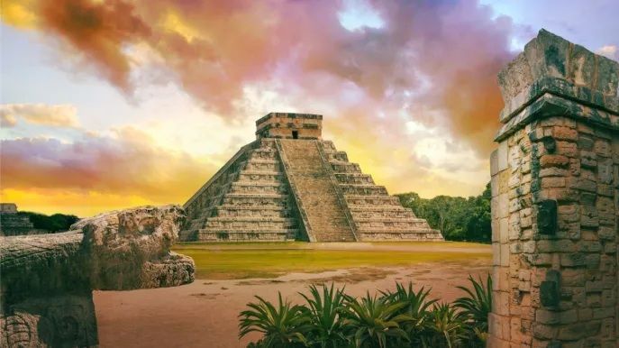 Un'imponente piramide Maya a gradoni in un sito archeologico, circondata da rovine di pietra e vegetazione sotto un cielo colorato al tramonto.