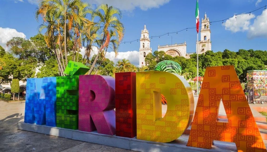 Grandi lettere colorate che compongono la parola 'MERIDA' si ergono in una piazza con una cattedrale, palme e una bandiera messicana sullo sfondo.