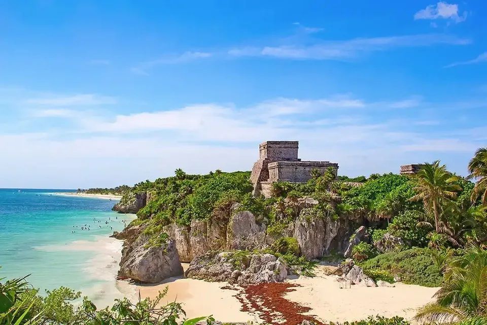 Antiche rovine in pietra si ergono su una lussureggiante scogliera che si affaccia su una spiaggia sabbiosa con acque turchesi sotto un cielo azzurro.