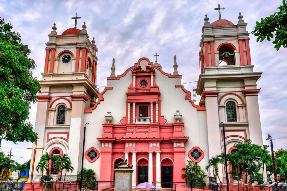 Symmetrische Fassade einer reich verzierten Kirche mit rosa-weißen Mauern, zwei Glockentürmen und Kreuzen vor bewölktem Himmel.