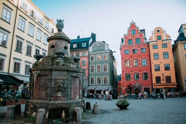 An ornate stone fountain stands in a cobblestone square, with colorful historic buildings in the background under a clear sky.