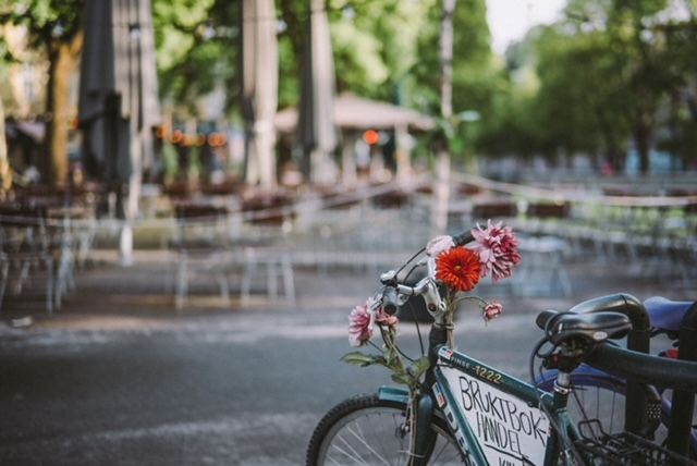 A close-up of a bicycle's handlebars decorated with red and pink flowers, with a blurry outdoor cafe in the background.