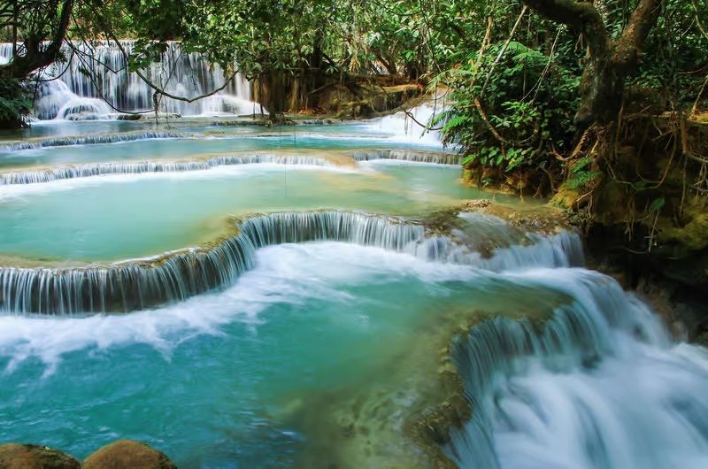 Foto a lunga esposizione di cascate a gradoni con acqua turchese in una giungla lussureggiante.