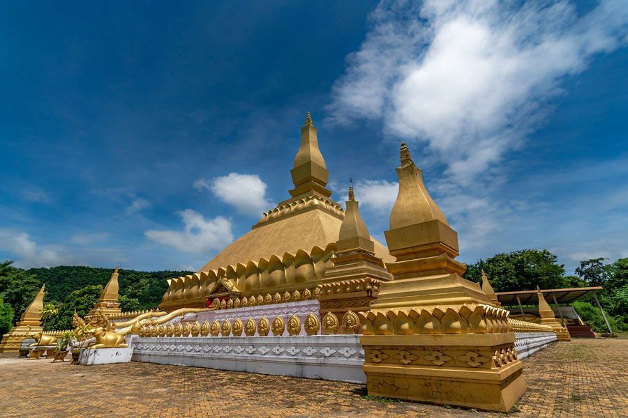 Una grande stupa dorata e riccamente decorata, con molteplici guglie, si erge in un cortile pavimentato sotto un cielo azzurro con nuvole.
