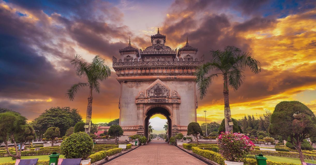 Al termine di un sentiero, in un parco con palme, un grande e ornato monumento ad arco si staglia contro un cielo colorato al tramonto.