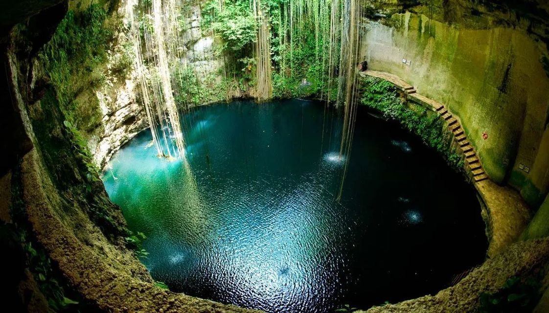 Una veduta grandangolare dall'alto di un grande cenote a cielo aperto con acqua blu profonda, circondato da pareti rocciose e piante rampicanti pendenti.