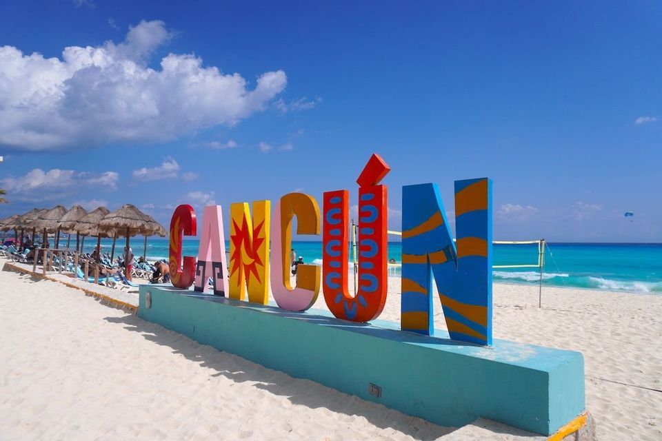 Un grande cartello colorato con la scritta 'Cancún' si trova su una spiaggia di sabbia bianca accanto all'oceano turchese sotto un cielo azzurro brillante.