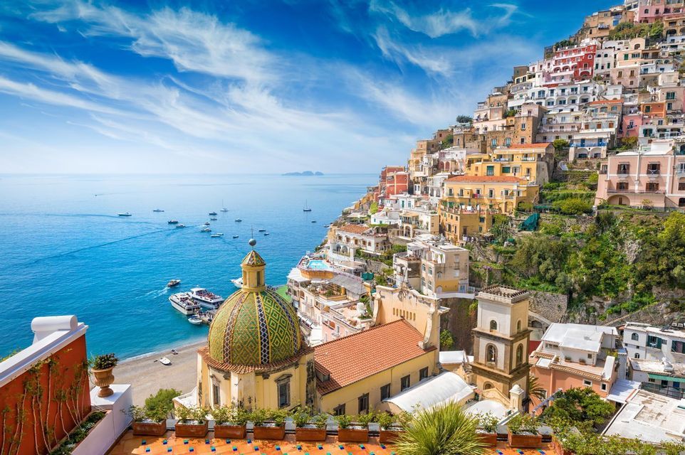 A colorful town built into a steep hillside with a tiled dome, overlooking a bay with boats under a blue sky.