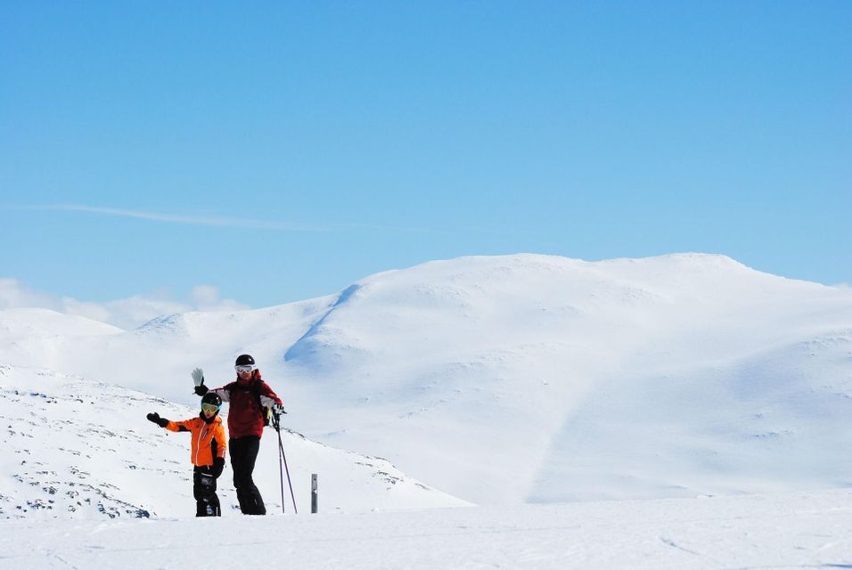 Due persone di un gruppo WeRoad in tenuta da sci che salutano su una montagna innevata sotto un cielo azzurro.