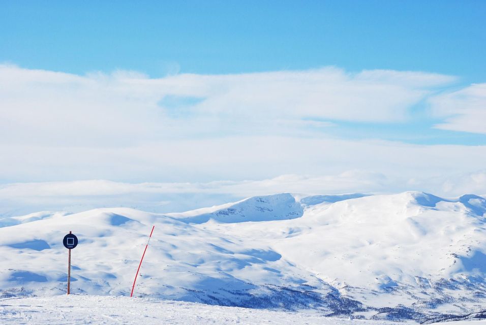 Un segnavia sulla pista da sci si staglia su un pendio innevato, affacciato su una vasta catena montuosa coperta di neve sotto un cielo azzurro.