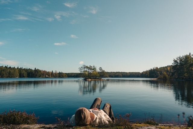 A person lies on a rocky shore, looking out over a calm lake with a small, tree-covered island in the distance.