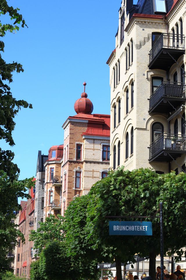 Ornate European buildings with a red turret stand along a street with leafy green trees and a sign reading 'BRUNCHOTEKET'.