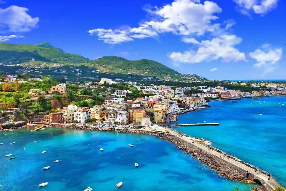 A vibrant coastal town on a green hillside with a stone pier jutting into a turquoise bay filled with small boats.