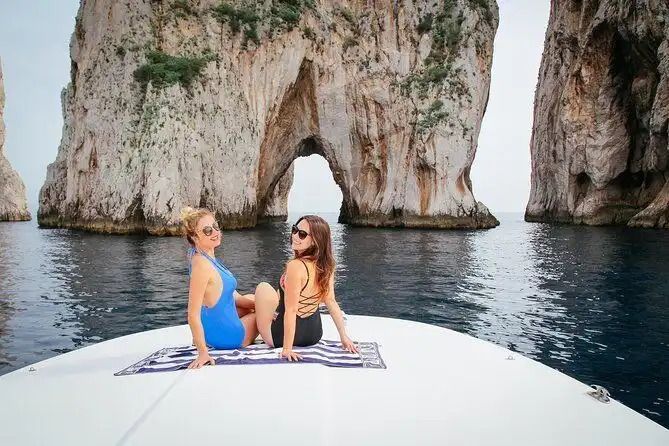 Two women in swimsuits sit on the bow of a boat, posing in front of large rock formations with a natural arch.