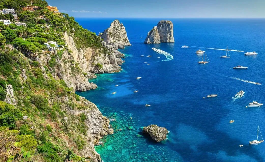 An aerial view of a rugged coastline with large rock formations and several boats sailing on the clear blue sea.