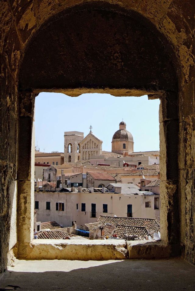 A historic city skyline with a church bell tower and dome, viewed through a rough, stone arched window under a clear sky.