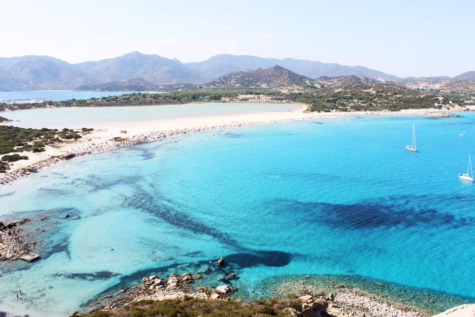 An aerial view of a sandy isthmus beach with turquoise water on both sides, with people swimming and two sailboats floating in the sea.