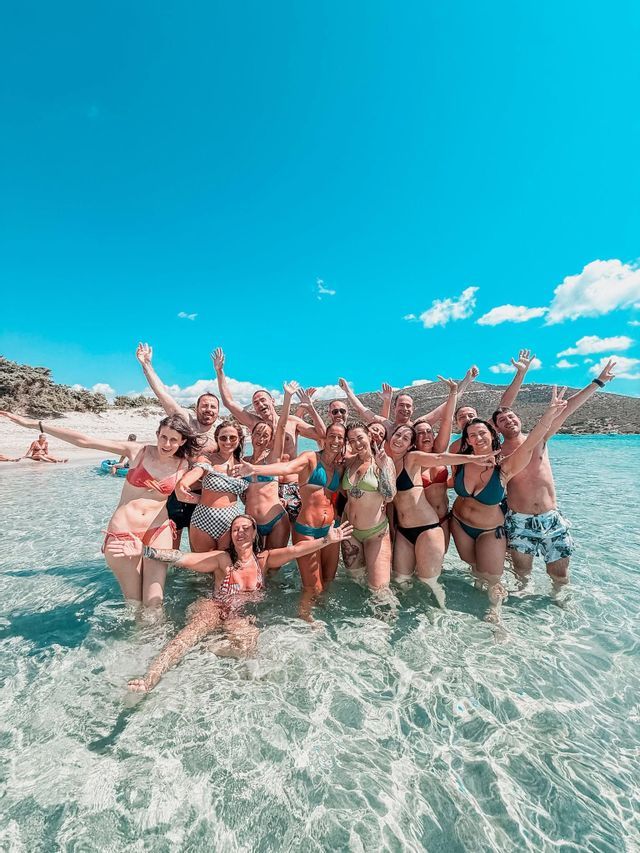 A WeRoad group trip of smiling people in swimwear posing together in the clear shallow water at a beach.