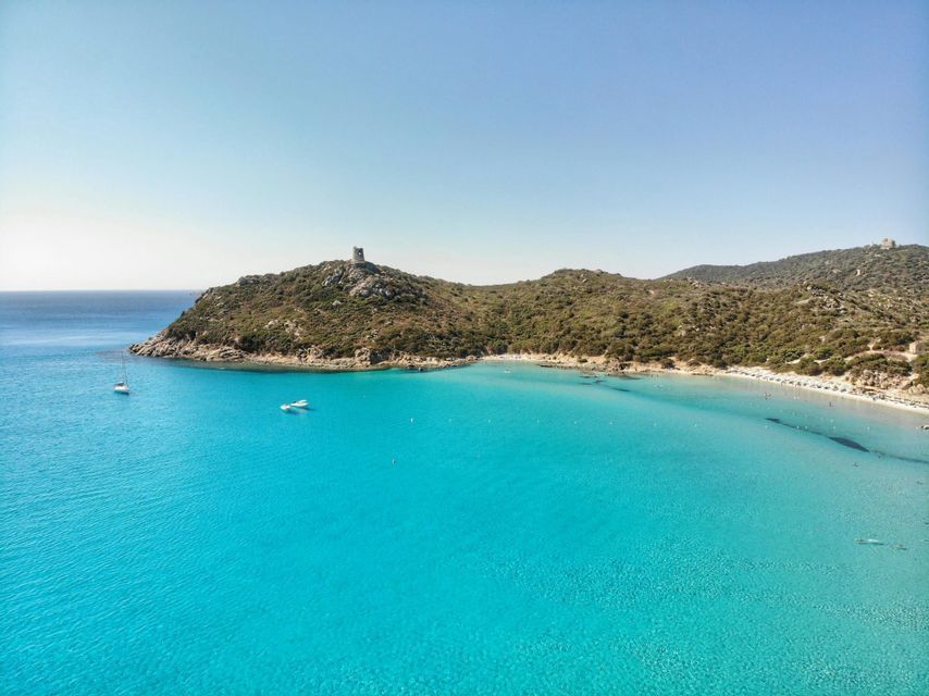 An aerial view of a bay with clear turquoise water, a sandy beach, and a green hill topped by a stone tower.