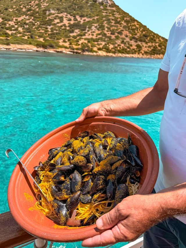A person holds a large terracotta bowl of spaghetti with mussels, topped with yellow seasoning, over turquoise water.
