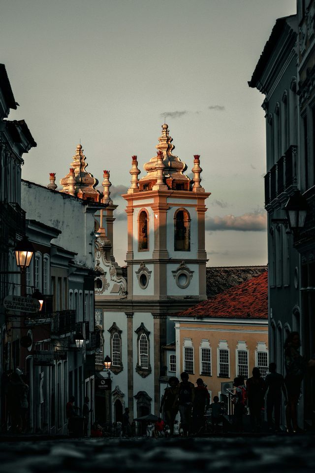Una chiesa sontuosa con due campanili, illuminata dal tramonto, vista da una strada di ciottoli in ombra sottostante.