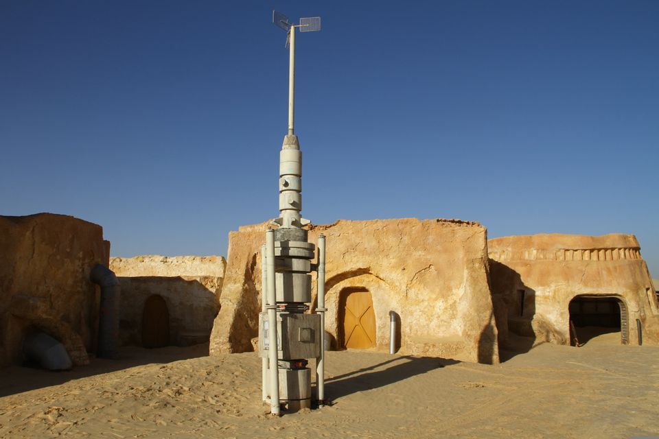 Ein weißer, futuristischer Turm steht inmitten sandfarbener Lehmgebäude in einer Wüstenlandschaft unter klarem blauem Himmel.