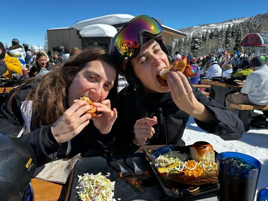 Dos mujeres de un viaje en grupo de WeRoad comen bocadillos en una mesa al aire libre en una estación de esquí soleada y nevada.
