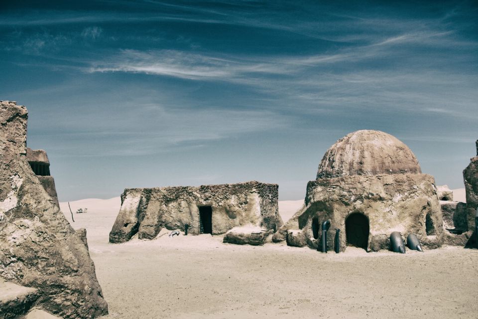 Sandfarbene Gebäude mit Kuppeldach stehen in einer weiten Wüste unter einem gestreiften blauen Himmel.