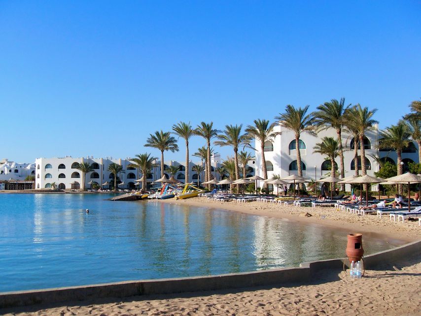 Ein Sandstrand vor weißen Hotelgebäuden, gesäumt von Palmen und Sonnenliegen, mit ruhigem Wasser unter klarem blauem Himmel.