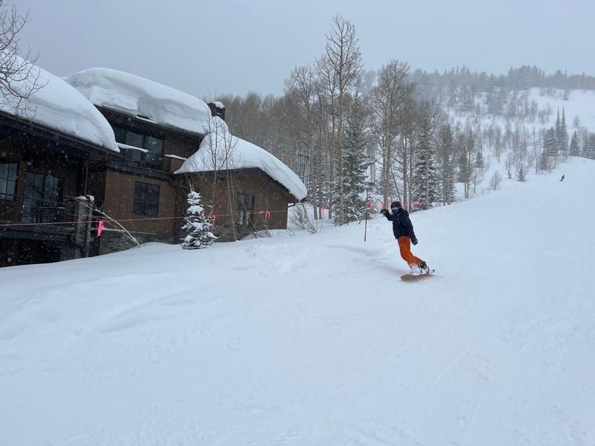 Una persona con chaqueta oscura y pantalones anaranjados hace snowboard en una pendiente nevada junto a casas y árboles cubiertos de nieve.