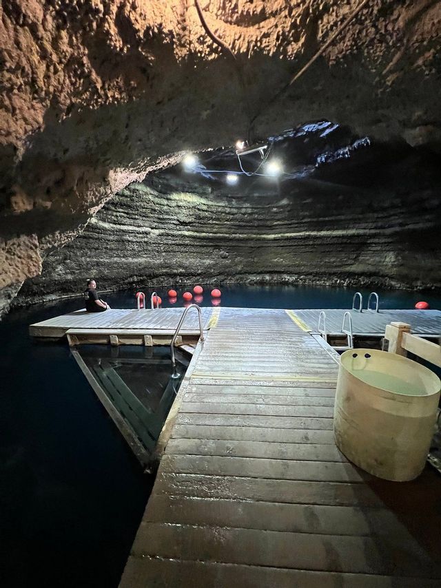 Una persona se sienta en un muelle de madera sobre agua azul oscura dentro de una cueva grande e iluminada.