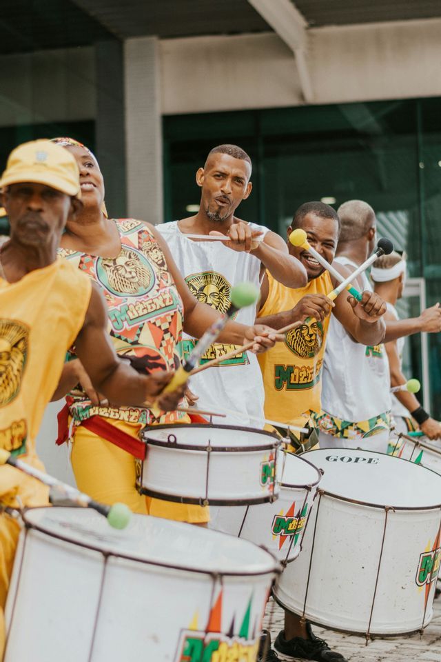 Un gruppo di musicisti in abiti coordinati gialli e bianchi suona grandi tamburi bianchi durante uno spettacolo all'aperto.