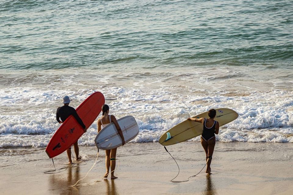 Un viaggio di gruppo WeRoad con tre surfisti che, tavole in mano, si avviano verso le dolci onde dell'oceano da una spiaggia di sabbia.