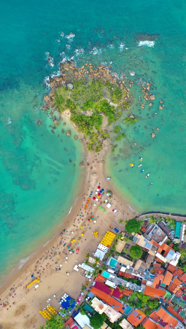Una vista aerea dall'alto di una lingua di sabbia che collega una città costiera a un isolotto roccioso e verde in acqua turchese, con persone e ombrelloni colorati sulla spiaggia.