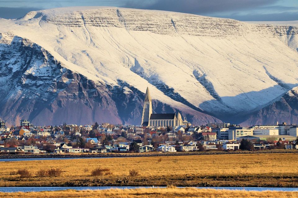 Una città con una chiesa imponente si trova ai piedi di una massiccia montagna innevata sotto un sole splendente.