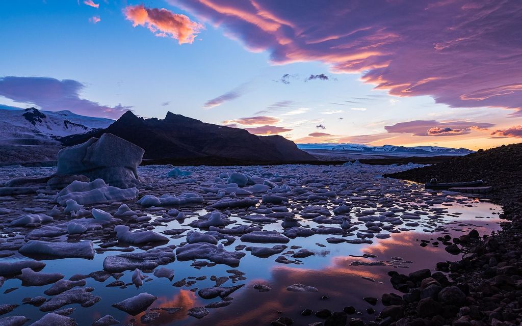 Iceberg galleggiano in un lago glaciale al tramonto con montagne sullo sfondo e nuvole colorate che si riflettono sull'acqua.