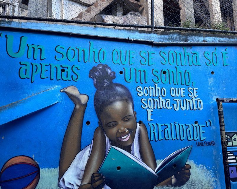 Un murale colorato su una parete blu brillante raffigura una bambina sorridente sdraiata che legge un libro, circondata da una citazione in portoghese.