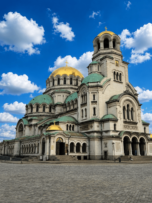 Une grande cathédrale blanche avec des dômes dorés et verts se dresse sur une place pavée sous un ciel bleu avec des nuages.