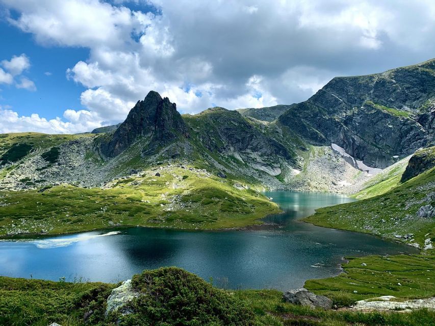 Un lac de montagne aux eaux claires et bleues est niché dans une vallée de montagnes verdoyantes et rocheuses sous un ciel partiellement nuageux.