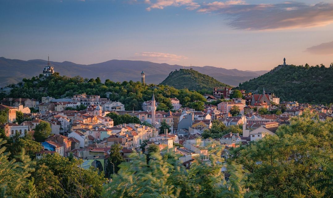 Une ville aux bâtiments colorés et aux toits de tuiles est nichée au milieu de collines verdoyantes sous un ciel de coucher de soleil.